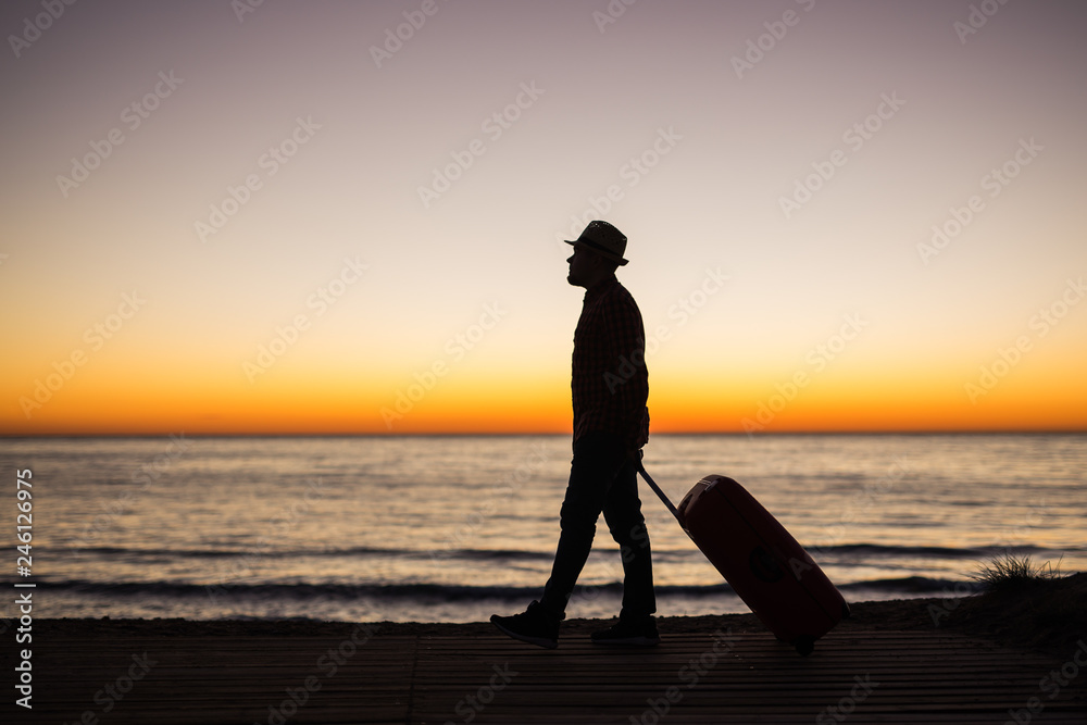 Vacation, summer and travel concept - young man silhouette with suitcase at sunset near the sea