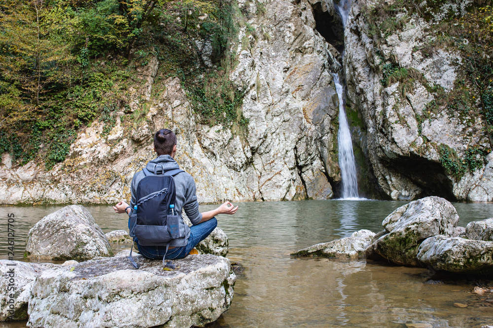 Young guy tourist in casual clothes with backpack sits in picturesque place on stone overlooking waterfall and meditates alone. Concept of self-knowledge of self-improvement and self-identification