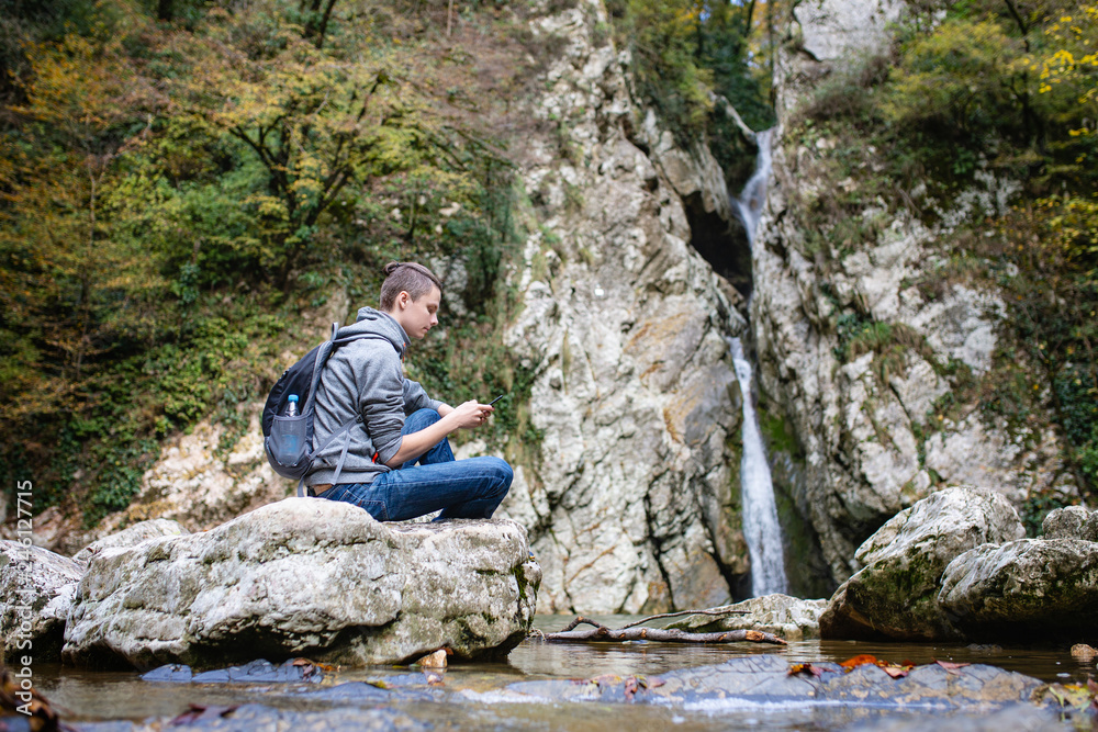 Side view - lonely tourist guy with backpack sits in picturesque place on stone by mountain river and communicates in social networks and smartphone. Concept of unity of modern technology and wildlife