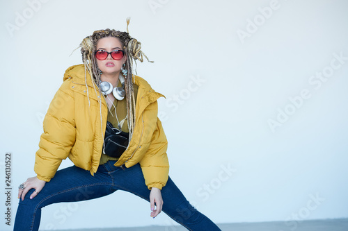 young blonde girl in sunglasses with african pigtails listens to music in headphones and dances against a white background. © yavdat