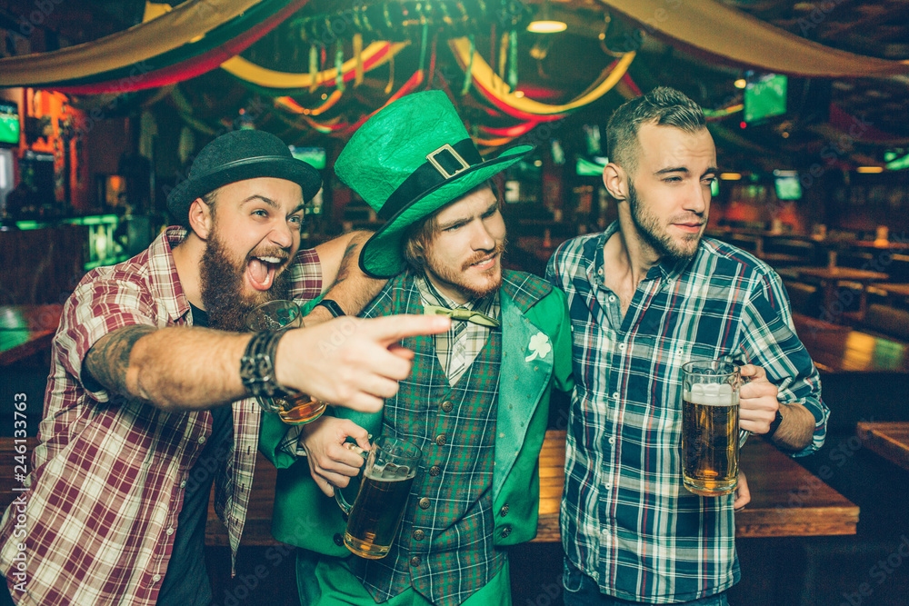 Excited young men stand in pub together. Guy on left point. They look to right. Young man in green suit wear St. Patrick's costume.