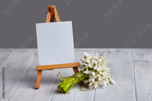 Bouquet of snowdrops and a small easel with a white paper and mini jars on agrey background. Greeting card with a space for text photo