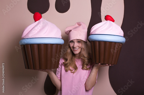 Excited blonde woman wearing pink cap enjoying big cupcakes at studio photo