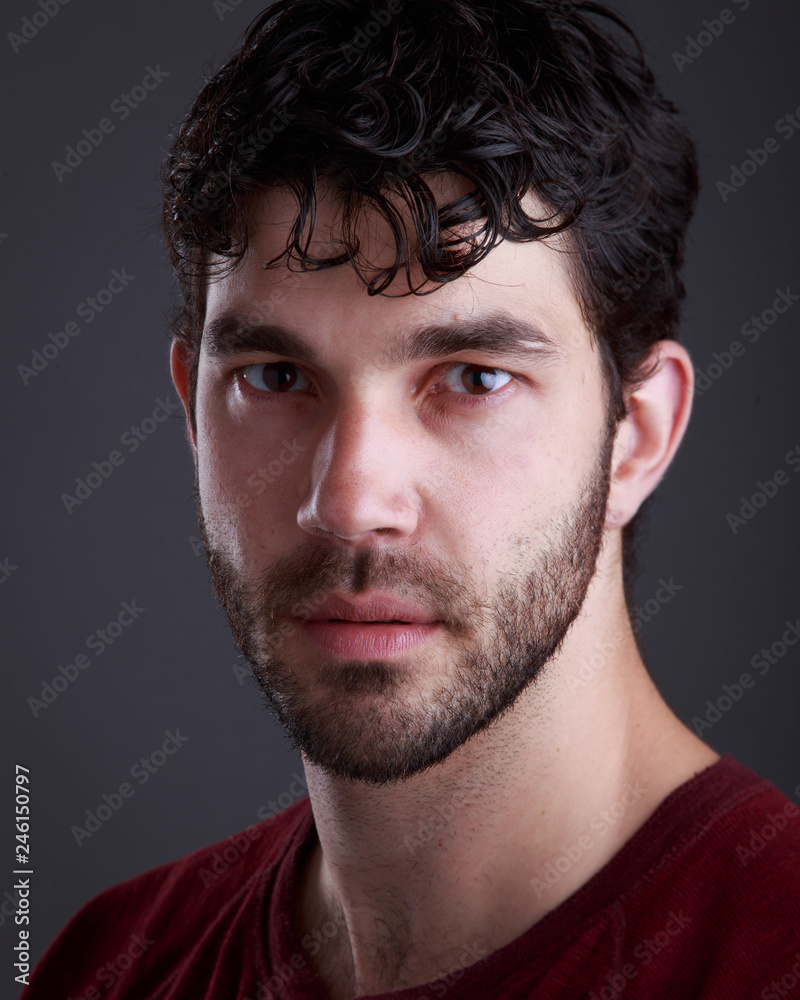 Fototapeta premium Headshot of handsome caucasian man with stubble and black hair and burgundy t shirt on grey background