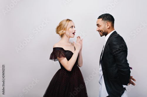 Lovely sweathearts of attractive astonished young woman in luxury evening dress and handsome man in tuxedo dating on white background. Valentines Day, surprise, true emotions, gift, love, together