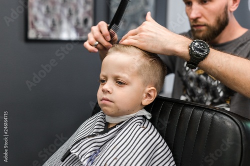 Serious little blond boy with blue eyes sitting on a chair in the Barber shop. Hairdresser cuts hair on the head of the child making a fashionable hairstyle