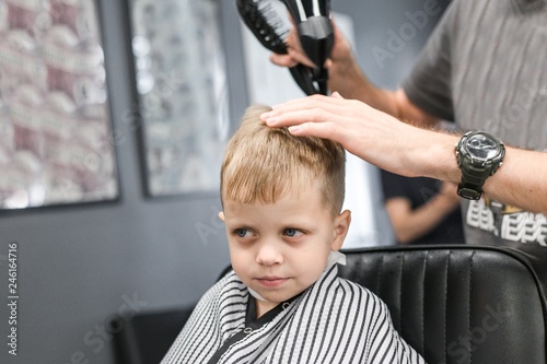 Little blond boy with blue eyes sitting on a chair in the Barber shop. Hairdresser cuts hair on the head of the child making a fashionable hairstyle