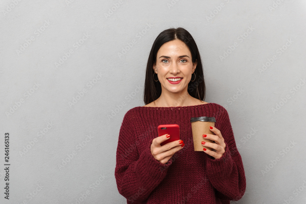 Pretty young woman dressed in burgundy sweater using mobile phone isolated over grey wall background. Pretty young woman dressed in burgundy sweater using mobile phone isolated over grey wall background.