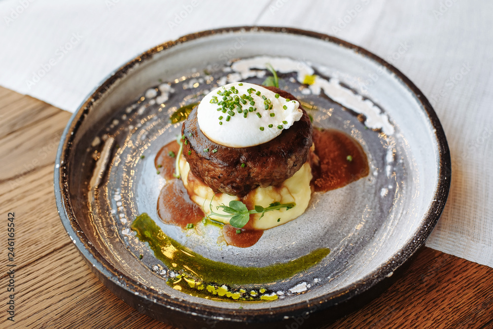  Meat cutlet on a pillow of mashed potatoes with poached egg on top, poured with pesto and teriyaki with meat sauce, decorated with green onions.
