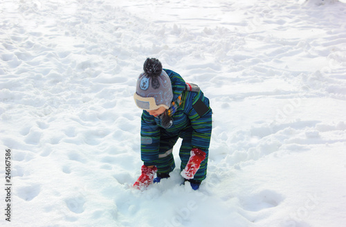A four-year-old child, a boy dressed in colorful winter clothes, plays with snow on a snowy ground on a sunny winter day.