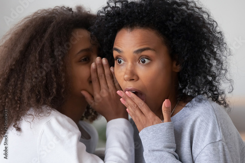 African American teenage girl tell secret to surprised young mom or nanny, teen daughter share gossip whispering in ear to shocked black mother, young parent and kid spend time together talking photo