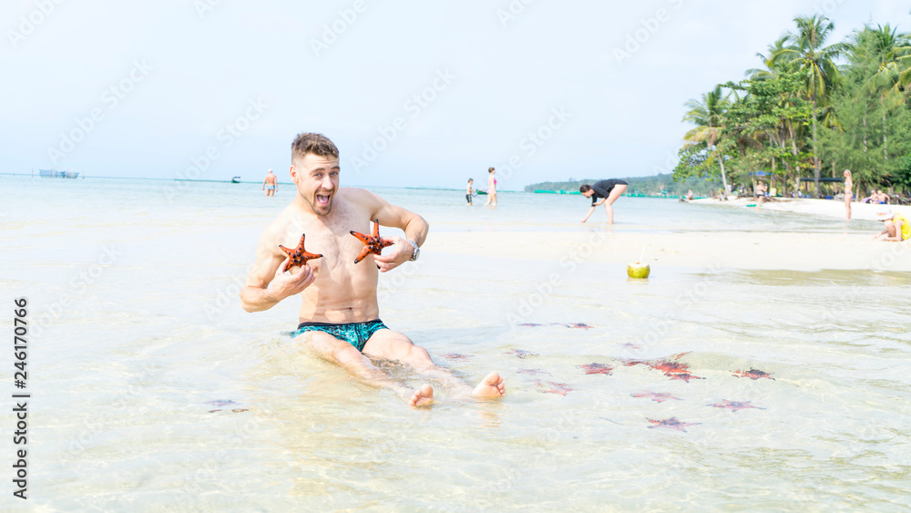 Travel boy is sitting on the sea with red starfishes makes funny photos in Phu Quoc island in crystal clear water