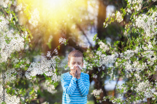 Boy sneezes in the park against the background of a flowering tree because he is allergic .