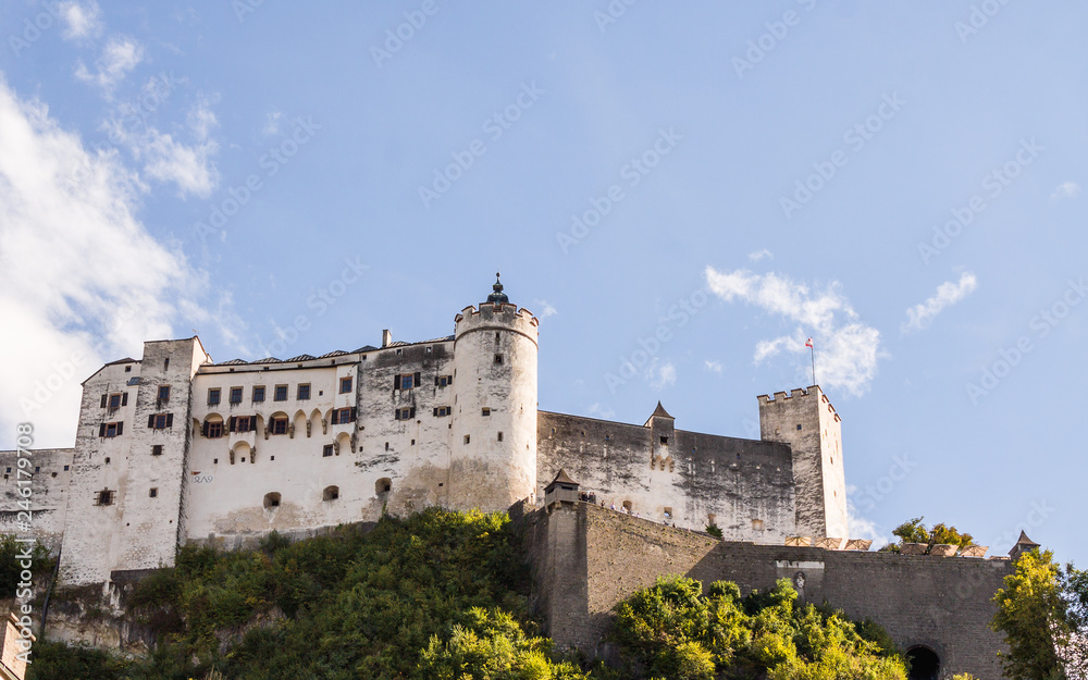 Burg auf Berg mit weisser Burgmauer und Türmen vor blauem Himmel mit Wolken