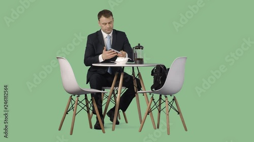 A man in a business suit sitting at a coffee table and drinking a tea. Transparent green screen background