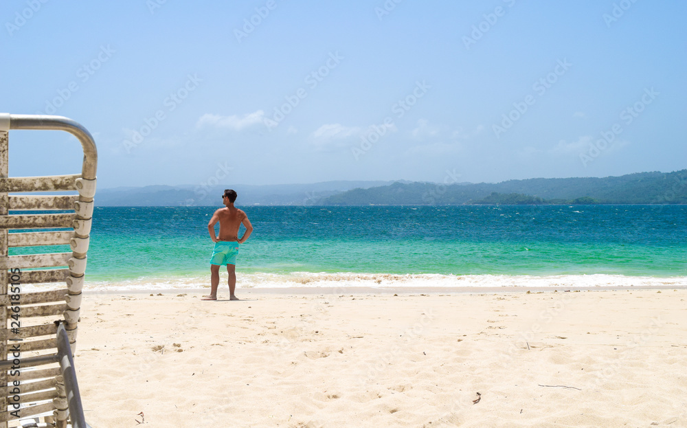  Man standing on the white beach in front of the ocean, sun lounger in front