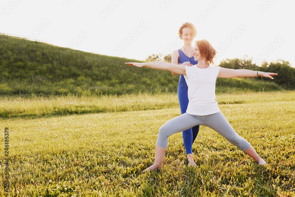 Pretty woman in a blue suit a yoga instructor helps her young woman client to do the yoga asana hero pose on a green lawn in a sunny summer day park. Healthy body concept Pretty woman in a blue suit a yoga instructor helps her young woman client to do the yoga asana hero pose on a green lawn in a sunny summer day park. Healthy body concept