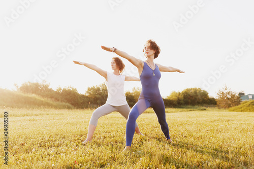 Two beautiful slim healthy young women in sportswear doing the pose of a hero yoga asana on a green lawn in a park on a sunny summer day. Healthy slim body concept. Advertising space © Rithor