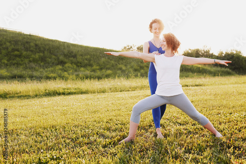 Pretty woman in a blue suit a yoga instructor helps her young woman client to do the yoga asana hero pose on a green lawn in a sunny summer day park. Healthy body concept