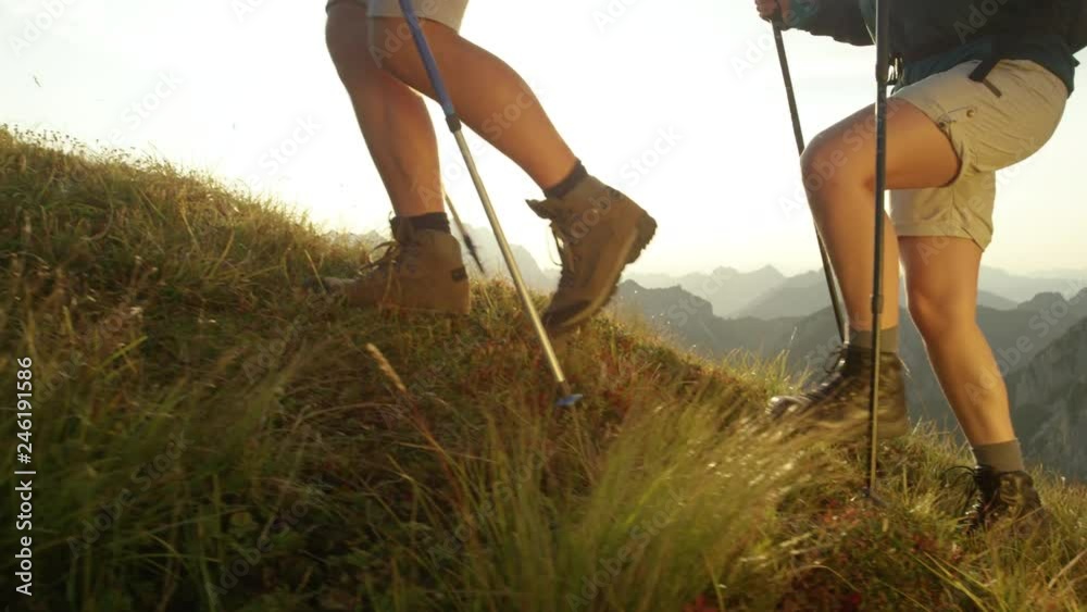 SLOW MOTION SUN FLARE CLOSE UP: Unrecognizable tourist couple on summer vacation exploring the scenic mountains on a sunny afternoon. Hikers wearing brown leather boots walking up a grassy hill in