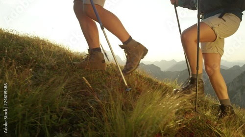 SLOW MOTION SUN FLARE CLOSE UP: Unrecognizable tourist couple on summer vacation exploring the scenic mountains on a sunny afternoon. Hikers wearing brown leather boots walking up a grassy hill in