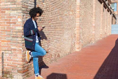 Canvas-taulu Young handsome African man using his smartphone with smile while leaning on a br
