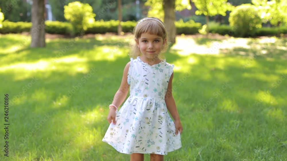A cute little girl in a summer dress whirls around herself in the park. Portrait of cute smiling little girl in Park on green grass background. A cute little girl in a summer dress whirls around herself in the park. Portrait of cute smiling little girl in Park on green grass background.