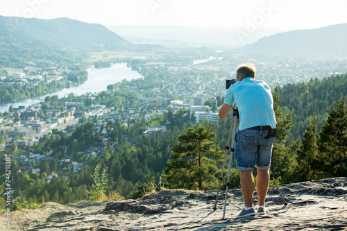 Man filming on camera with tripod on the top of mountain. Tourist hiking and taking pictures. Panoramic top view of Drammen city, mountains in fog and valley with river, sunny weather. Norway