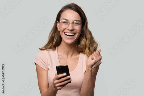 Happy funny millennial woman euphoric celebrating mobile win victory triumph holding phone, cheerful excited girl laughing having fun using cellphone apps isolated on white studio blank background