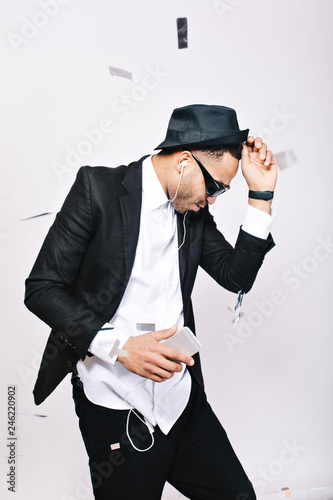 Fashionable excited handsome guy in suit, hat, black sunglasses having fun in tinsels on white background. Listening to music through headhones, dancing, celebrating party, cool