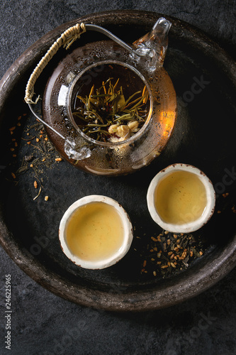 Hot green tea in two traditional chinese clay ceramic cup and opened glass teapot in tray over black marble background. Flat lay, close up