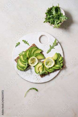 Vegan sandwiches with sliced avocado and lemon on rye bread, arugula salad served on ceramic board over white marble background. Flat lay, space