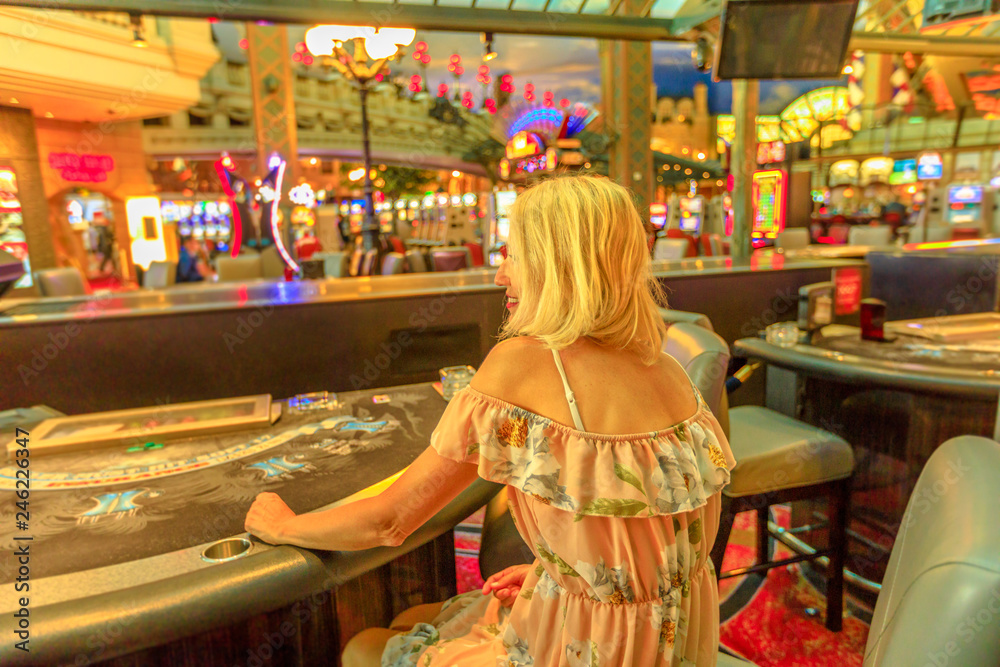 Back side of caucasian blonde woman gambling in casino with bright lights. Lifestyle girl waiting to play at blackjack in Las Vegas, Nevada, United States. Gambler addict concept