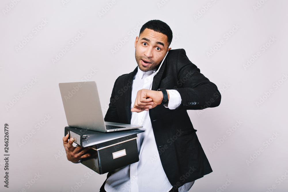 Busy hard-working guy in white shirt and black jacket talking on phone, holding folders, laptop, looks astonished at watch on white background. Office worker, modern technology, career Busy hard-working guy in white shirt and black jacket talking on phone, holding folders, laptop, looks astonished at watch on white background. Office worker, modern technology, career