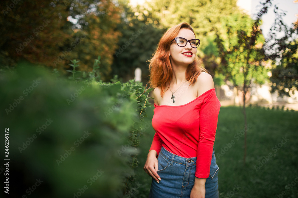 Attractive redhaired woman in eyeglasses, wear on red blouse and jeans skirt posing at green park. Attractive redhaired woman in eyeglasses, wear on red blouse and jeans skirt posing at green park.
