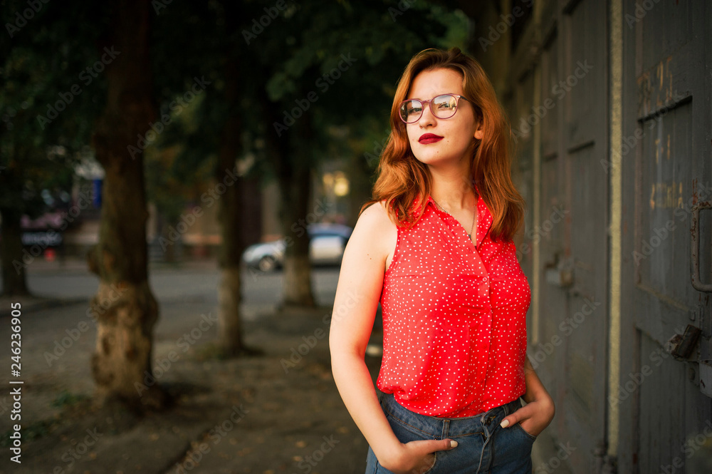 Attractive redhaired woman in eyeglasses, wear on red blouse and jeans skirt posing. Attractive redhaired woman in eyeglasses, wear on red blouse and jeans skirt posing.