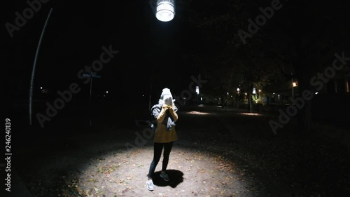 Girl with closed eyes playing hide-and-seek in dark street. Young woman in warm clothes in park. Streetlight. Dangerous entertainment. Evening time in big city.