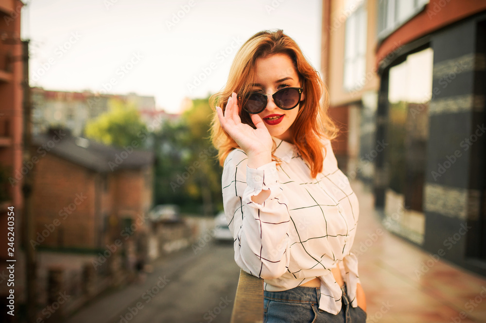 Fototapeta premium Attractive redhaired woman in sunglasses, wear on white blouse posing at street against modern building.