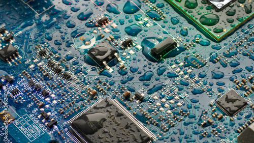The circuit board of the motherboard is covered with drops of water in blue-green tones. Close up computer part. 16:9 image, selective focus