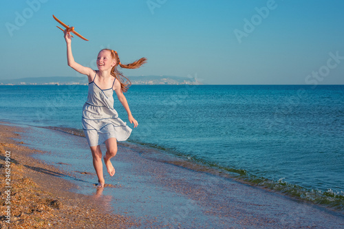 Seashore and girl. Happy red-haired child, playing with a toy airplane, runs along the sandy beach of the sea against the blue summer sky.
