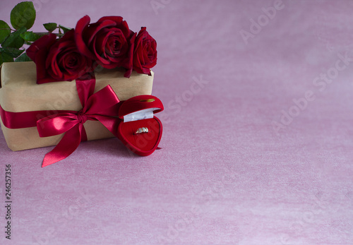 red box with engagement ring with precious stones, box tied with red ribbon, three red roses on pink background (horizontally, copy space)