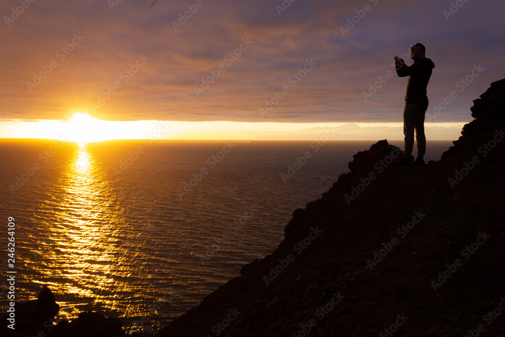 Photographer with cellphone standing on cliff edge rock capturing beautiful sunset by the sea in Gran Canaria. Silhouette of man taking photo with smartphone at twilight. Travel memories concept