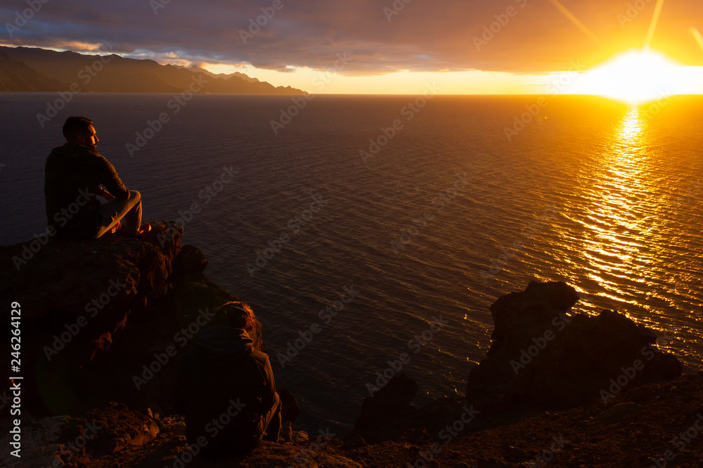 Couple of friends sitting on cliff edge rock staring at sunset by the sea in Gran Canaria, Spain. Young men contemplating twilight with sun reflecting on calm ocean. Travel destination concept