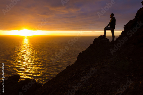 Hiker silhouette standing on cliff edge rock staring at sunset in Gran Canaria, Spain. Fearless man enjoys splendid twilight by the sea in Canary Islands. Visionary, adventure, challenge concepts photo