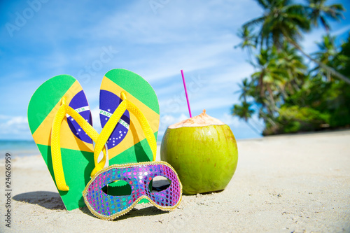 Brazil flag flip-flops and brightly sequined carnival mask resting together in the sand on the shore of a palm-fringed Brazilian beach 