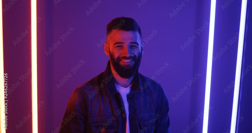 Handsome young Caucasian guy standing in the pink and blue neon lamps light, turning his head to the camera and smiling joyfully. Portrait shot. Handsome young Caucasian guy standing in the pink and blue neon lamps light, turning his head to the camera and smiling joyfully. Portrait shot.