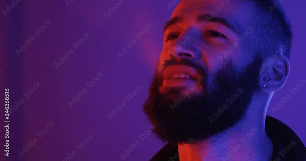 Close up of the attractive young Caucasian man with a beard smoking a cigarette in the neon pink and blue light. Portrait. Close up of the attractive young Caucasian man with a beard smoking a cigarette in the neon pink and blue light. Portrait.