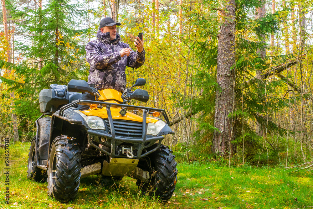 Fototapeta premium ATV. A man rides through the forest on an all-terrain vehicle. Quad bike. In the woods. Trip to the forest on ATV. The traveler checks the route on the map on the phone.