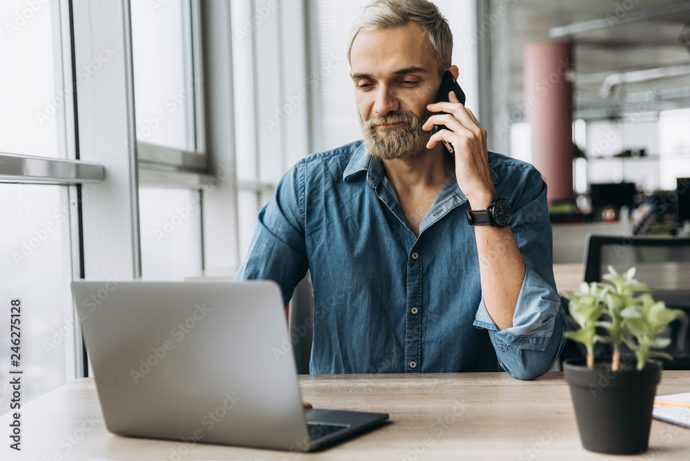 The guy works in a laptop. A very busy man talking on a smartphone during working process The guy works in a laptop. A very busy man talking on a smartphone during working process