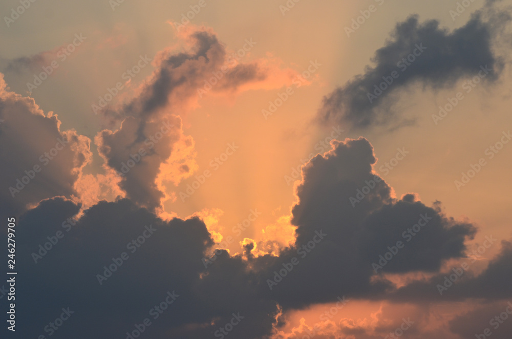 The rising sun is  hidden by morning clouds. The clouds range from dark grey to light orange in colour. Streams of sunlight spread out from behind the clouds into a pale sky.